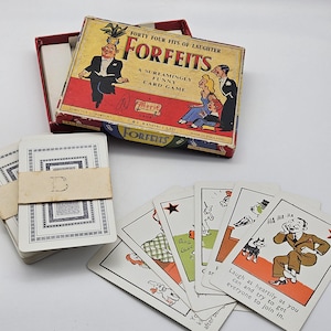 May include: A vintage Forfeits card game box with red and yellow graphics featuring cartoon characters, accompanied by a stack of playing cards bound with a paper band marked with the letter D and several loose cards with humorous illustrations.