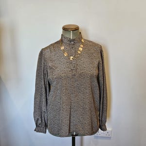 May include: A long-sleeved houndstooth print blouse in brown and black, displayed on a mannequin. The blouse has a high collar and a few buttons at the neckline. A gold-toned necklace with circular pendants adorns the blouse.