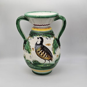 Può includere: Un vaso in ceramica dipinto a mano con due manici verdi. Il vaso presenta uno sfondo bianco con una dettagliata illustrazione di un uccello. Foglie e viti verdi decorano la parte superiore. Il vaso ha un bordo e una base verdi, con accenti gialli e rossi.