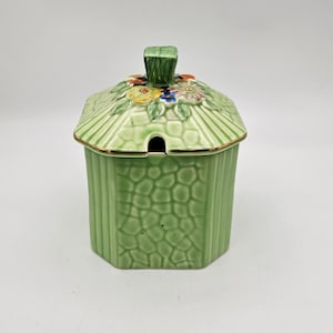 May include: A light green ceramic sugar bowl with a lid. The lid is decorated with colourful flowers and a green handle. The body of the bowl has a textured pattern. The rim of the lid is trimmed with gold.