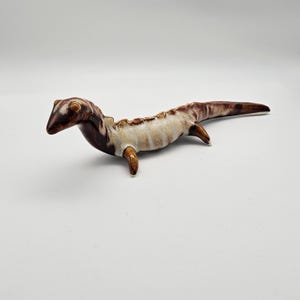 Può includere: Una statuetta di lucertola in ceramica con smalto marrone e bianco. La lucertola è in posizione di strisciamento con la coda estesa.