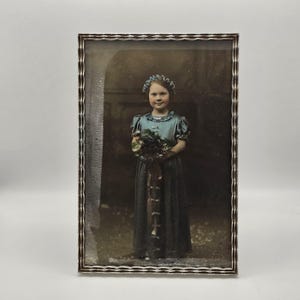 Puede incluir: Fotografía antigua enmarcada de una niña con un vestido azul sosteniendo un ramo. El marco plateado tiene un diseño texturizado y acanalado. La niña lleva un tocado floral y la fotografía tiene un tono sepia.