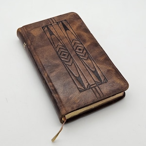 May include: A brown leather book with an embossed geometric design on the cover. The book has a worn appearance and a gold-coloured page edge. A thin, tan ribbon bookmark extends from the book's spine.