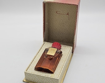May include: A small amber-colored glass perfume bottle with a red cap, featuring a gold label with the text Coty, presented in an open rectangular box with a red exterior and a cream-colored interior patterned with small floral motifs.