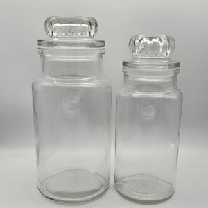 May include: Two clear glass jars with lids. The jars are of different sizes and have a rounded rectangular shape. The lids have a raised, textured design.
