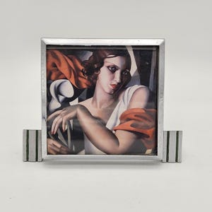 May include: A silver picture frame with a square portrait of a woman in a white dress and red shawl. The frame has a rectangular base with green stripes. The artwork features a woman with red lipstick and dark hair.