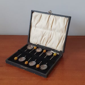 May include: A black velvet-lined box with a hinged lid, containing a set of eight silver teaspoons with amber-colored glass handles. The spoons are arranged in rows, with the handles facing up.