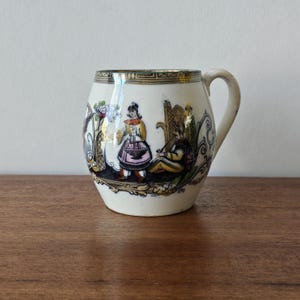 Op de afbeelding: Een kleine, gebroken witte keramische mok met een gebogen handvat. De mok heeft een kleurrijke illustratie van twee figuren in kleding uit een bepaalde periode, met bloemen- en geometrische patronen. De rand heeft een groene en gouden rand.