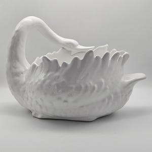 May include: A white ceramic swan-shaped bowl. The swan has a long, curved neck and a detailed feather design. The bowl is designed to hold items and is a decorative piece.