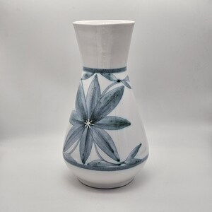 May include: White ceramic vase with a flared top and a rounded body. The vase is decorated with a large blue floral design and a blue band around the neck. The floral design has a star-like centre.