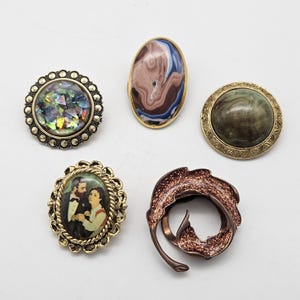 Op de afbeelding: Een verzameling van vijf decoratieve spelden en broches. Eén heeft iriserende fragmenten in een kralenframe, een andere een bruin en blauw gemarmerd ovaal, een derde een groen gemarmerde cirkel, één toont een portret van een koppel en één is een koperen blad.