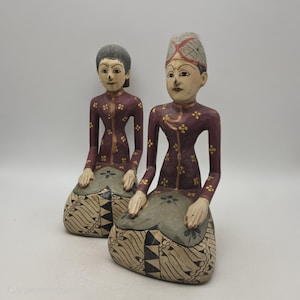 Könnte beinhalten: Zwei handgeschnitzte Holzfiguren, vermutlich aus Indonesien, in traditioneller Kleidung. Die Figuren haben weinrote Oberteile mit goldenen Blumenmustern und gemusterte Röcke. Eine Figur trägt einen hohen Hut mit einem rot-weißen Design.