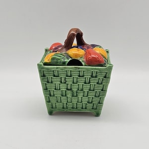 May include: A square ceramic sugar bowl designed to look like a green woven basket. The lid features a sculpted brown handle and a colourful assortment of raised fruit including red strawberries, yellow lemons, and purple grapes.