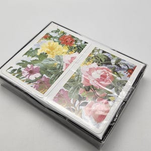 May include: A sealed deck of playing cards featuring floral designs. The cards display colorful illustrations of roses, petunias, and other flowers. The deck is encased in clear plastic and has a black border.