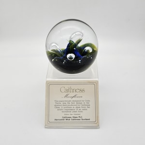 Puede incluir: Un pisapapeles esférico de cristal transparente con un diseño abstracto azul y verde. El pisapapeles se encuentra sobre un soporte de plástico transparente. El texto "Caithness Moonflower" es visible en una tarjeta debajo.