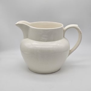 以下が含まれることがあります： 丸みを帯びたボディ、注ぎ口、湾曲したハンドルを備えたオフホワイトのセラミックピッチャー。ピッチャーは滑らかで光沢のある仕上がりで、クラシックなデザインです。ヴィンテージスタイルのキッチン用品アイテム。