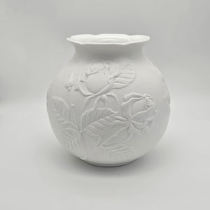 May include: White ceramic vase with a round body and a scalloped rim. The vase features embossed rose designs with detailed leaves and flower buds. The floral pattern is repeated around the vase, creating a textured surface.