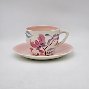 May include: A vintage teacup and saucer set. The cup and saucer are a soft pink colour. The cup features a floral design in shades of pink, purple, and blue. The interior of the cup is also pink. The set is likely made of ceramic.