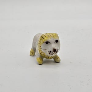 Pode incluir: Pequena estatueta de leão em cerâmica. O leão tem um corpo branco com detalhes amarelos na juba, patas e orelhas. O rosto apresenta pontos pretos para os bigodes e um nariz castanho. A estatueta está em posição agachada.