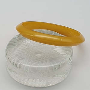 May include: A yellow plastic bangle bracelet. The bracelet is smooth and shiny. It is sitting on a clear glass surface.