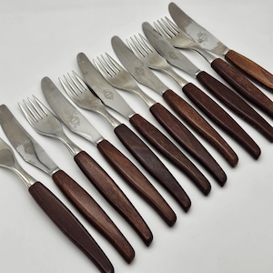 May include: A set of vintage silverware, including forks and knives, with stainless steel blades and dark brown wooden handles. The utensils are arranged diagonally on a white surface, showcasing their design and craftsmanship.