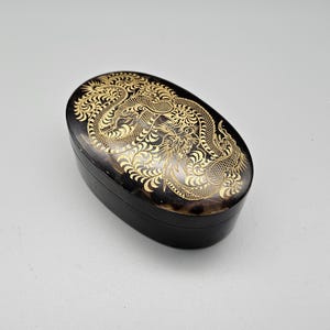 Könnte beinhalten: Eine kleine, ovale, schwarze Schachtel mit einem goldenen Drachenmotiv auf dem Deckel. Die Schachtel ist aus Holz und hat eine glänzende Oberfläche.