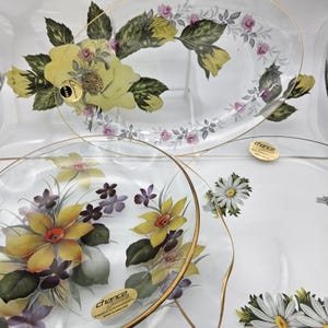 Puede incluir: Platos de servir de vidrio transparente con diseños florales. Los platos presentan flores amarillas, moradas y blancas con hojas verdes y bordes dorados. El plato ovalado tiene una gran flor amarilla en el centro.