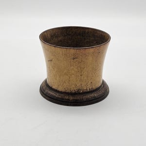 Pode incluir: Um pequeno recipiente cilíndrico com uma borda superior alargada e uma base circular larga. O objeto apresenta um acabamento desgastado com um corpo em tom dourado metálico e uma borda e base em castanho escuro, com textura visível.