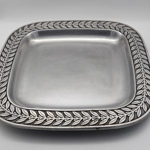 May include: A square, silver-coloured metal tray with a decorative laurel leaf border. The tray has a slightly recessed centre and rounded corners. The border features a detailed relief pattern of leaves and stems, adding a touch of elegance to the serving piece.