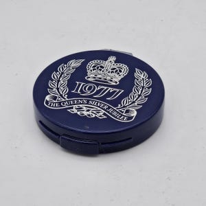 Könnte beinhalten: Ein marineblaues Kompaktetui mit einem weißen Emblem zum Gedenken an das silberne Thronjubiläum der Königin im Jahr 1977. Das Emblem enthält eine Krone, das Jahr 1977 und Lorbeerzweige. Das Kompaktetui hat einen Klappdeckel.