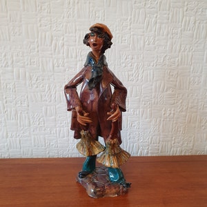 Puede incluir: Una figurita de cerámica de una persona que lleva un abrigo marrón, una bufanda azul y un sombrero marrón. La persona sostiene dos escobas en sus manos. La figurita está de pie sobre una base que parece adoquines.