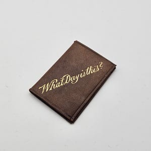 May include: A brown leather wallet with gold lettering that reads "What Day is this?"