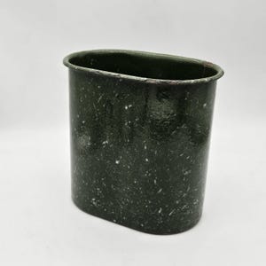 May include: A dark green, oval-shaped metal cup with a speckled pattern. The cup has a slightly worn rim and a smooth, cylindrical body. The overall aesthetic suggests a vintage or military-style design.