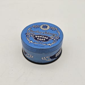 Può includere: Una scatola di latta blu vintage con la scritta "Huntley & Palmers Dundee Cake" in bianco. La scatola ha un design floreale e le parole "Reading & London" sono stampate in basso.