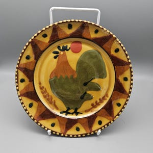 May include: A yellow ceramic plate featuring a brown, green and red rooster design. The plate has a brown and yellow border with black dots.