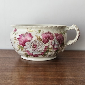 May include: Cream-colored ceramic bowl with a large handle and floral design. The bowl features a detailed pattern of pink and white flowers with green leaves. The handle has a decorative, ornate design.