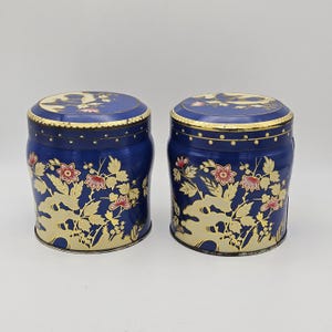 Puede incluir: Dos latas decorativas azules a juego con diseños florales dorados y flores rojas. Las tapas son azules con ribete dorado y un diseño de pájaro. Las latas son cilíndricas y tienen una estética vintage.