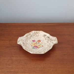 Puede incluir: Un plato de cerámica blanca con un borde dorado y un diseño floral. El plato tiene un borde festoneado y dos pequeñas asas.