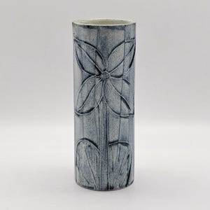 May include: A cylindrical ceramic vase with a mottled blue and grey finish. The exterior features a debossed floral design consisting of a four-petaled flower at the top and two large leaves at the base, connected by a vertical stem line.