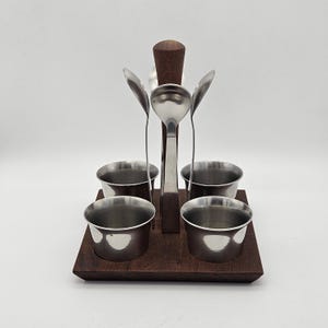 Könnte beinhalten: Ein Vintage-Gewürzset mit vier kleinen Edelstahlschalen und fünf Löffeln. Die Löffel und Schalen sind auf einem dunklen Holzsockel mit einem zentralen Holzgriff angeordnet. Ideal für Saucen oder Dips.