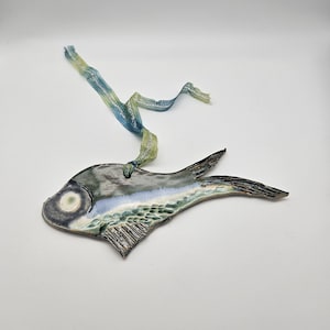 Puede incluir: Un adorno de pescado de cerámica con un esmalte azul, verde y blanco. El pez tiene un ojo blanco y un cuerpo texturizado. Está colgado de una cinta azul y verde.