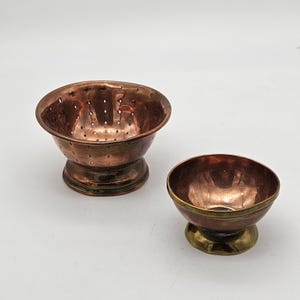 Könnte beinhalten: Zwei Kupferschalen mit Vintage-Ästhetik. Eine Schale ist ein Sieb mit kleinen Löchern, die andere eine kleinere Schale mit Messingfuß. Die Schalen haben einen warmen, rötlich-braunen Farbton.