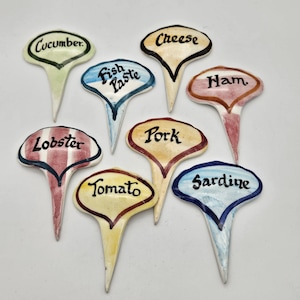 May include: A set of eight ceramic food markers with pointed bases. Each marker is a different colour and features a handwritten food label, including "Cucumber," "Fish Paste," "Cheese," "Ham," "Lobster," "Pork," "Tomato," and "Sardine."