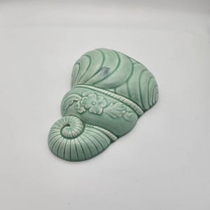 Puede incluir: Un aplique de pared de cerámica verde mar en forma de concha. La concha presenta un diseño floral y un detalle en espiral. Este artículo es decorativo.
