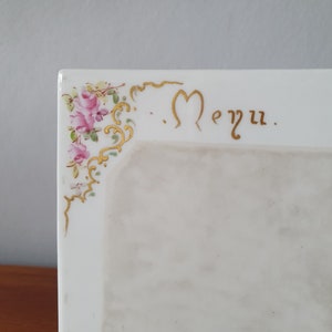 Può includere: Un tableau de menu in porcellana bianca con un bordo dorato e un motivo floreale. La parola "Menu" è scritta in caratteri dorati.