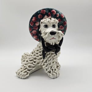 May include: White ceramic figurine of a dog wearing a green and pink hat with a black bow tie.