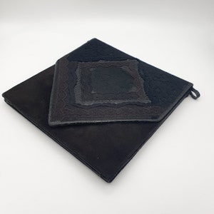 May include: A black suede clutch bag with a decorative flap featuring a layered, geometric design. The flap has a black, textured fabric with a diamond shape.