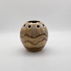 May include: A spherical ceramic flower frog vase featuring a glossy glaze in shades of tan, brown, and cream. The surface displays horizontal wavy bands of colour, and the top section contains multiple circular openings for arranging stems.