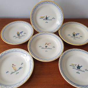 以下が含まれることがあります： 青と黄色の縁取りが施された白いセラミック製のプレート6枚セット。各プレートには、緑色の服を着た人物と鳥が手描きで描かれています。