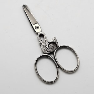 May include: Silver scissors with a swan-shaped handle. The scissors feature a detailed swan design with a red eye accent. The blades are straight, and the finger holes are oval.